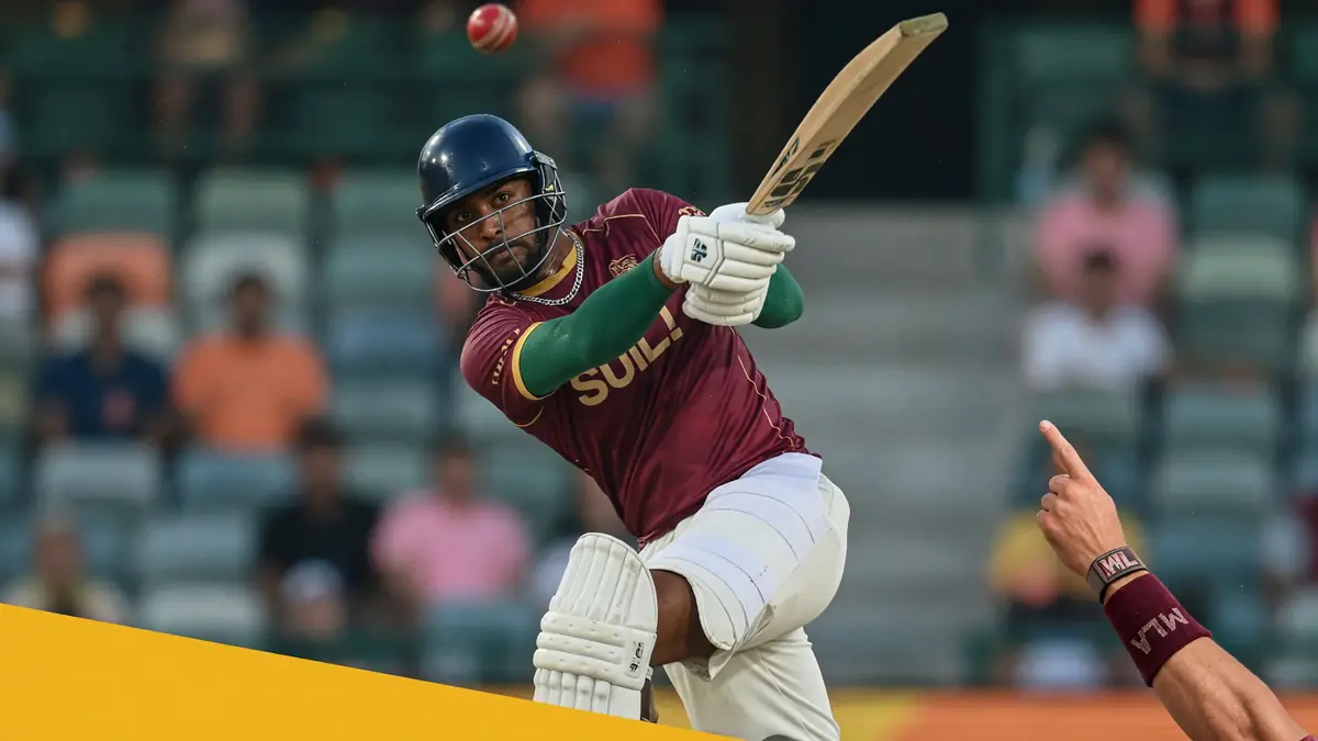 Maroon-kit batter in a blue helmet swinging hard as a red ball hangs above the bat under stadium lights