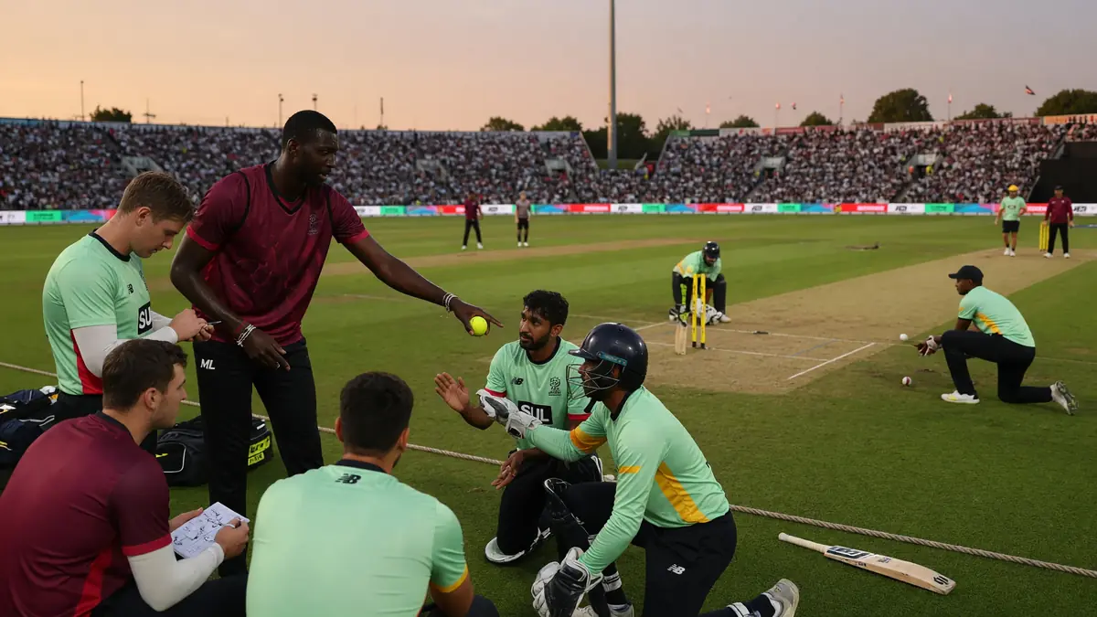 Mint-and-maroon players huddle on the outfield while a coach holds a yellow practice ball before a floodlit match